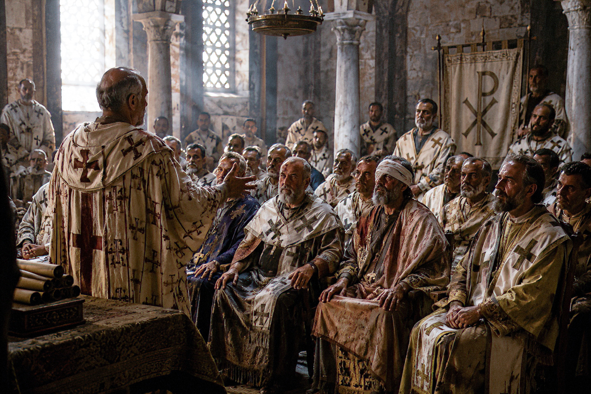 Clergy in cream robes with cross motifs listening to a speaker in a stone church interior